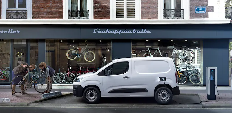 E-berlingo van blanc qui recharge sa batterie en ville devant un magasin de vélo
